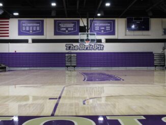 An indoor basketball court with an above bleacher sign titled “The Brit Pit.” The bleachers are purple and above a tan wood lacquered floor.