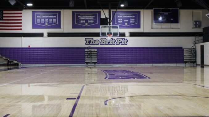 An indoor basketball court with an above bleacher sign titled “The Brit Pit.” The bleachers are purple and above a tan wood lacquered floor.