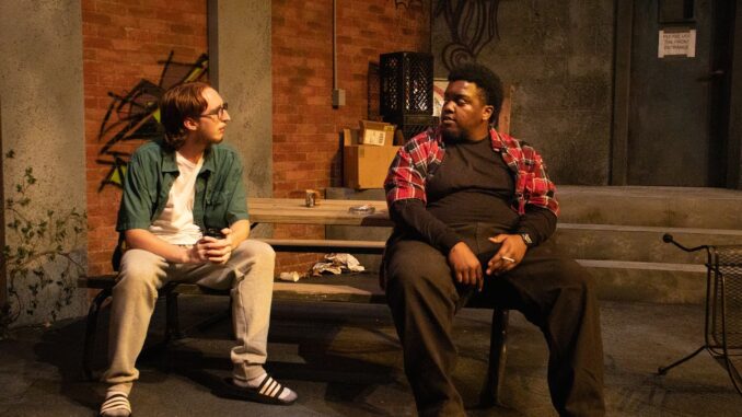 TheAliens Two male cast members sit next to each other on a wooden picnic table. They are both dressed in ordinary day clothes, one with a lighter color scheme than the other. The stage itself is modeled to represent a dingy alley behind a restaurant, complete with faux graffiti on the walls and miscellaneous brown cardboard boxes.