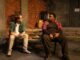 Two male cast members sit next to each other on a wooden picnic table. They are both dressed in ordinary day clothes, one with a lighter color scheme than the other. The stage itself is modeled to represent a dingy alley behind a restaurant, complete with faux graffiti on the walls and miscellaneous brown cardboard boxes.