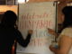 Two young adults with long hair in casual dress stand with their backs to the camera, writing with marker on a large, vertical white paper sign that faces the camera. The sign reads “Celebrate Hispanic Heritage Month with OLA” in colorful block letters.