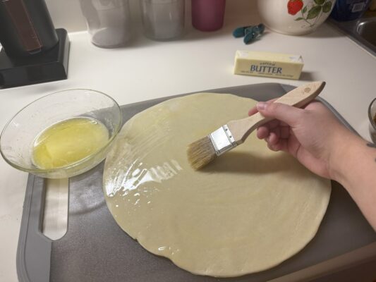 A bowl of melted butter and pie crust sits atop a cutting board. A hand with a brush spreads melted butter onto the pie crust. In the background is a stick of butter and a chip clip.