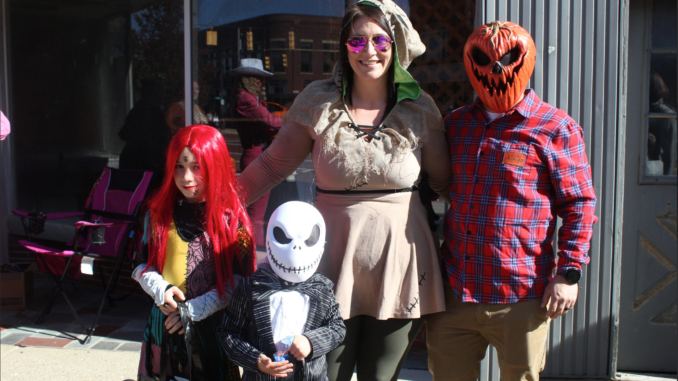 1st image A family dressed as popular Halloween movie characters, Sally, Jack, Oogie Boogie and The Pumpkin King from “The Nightmare Before Christmas” stand outside a building. Behind them are large glass windows.