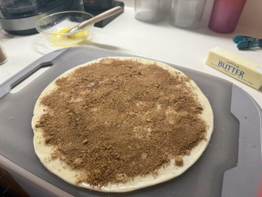 Pie crust with cinnamon and brown sugar lays flat atop a cutting board. In the background is a bowl with melted butter and a brush alongside a stick of butter.