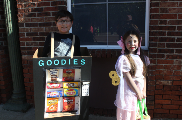 A young boy dressed in a cardboard box with pictures of vending machine snacks attached and a sign reading “GOODIES” stands next to a young girl in a pink dress with a gold wind-up piece attached to the back and cracked face paint.