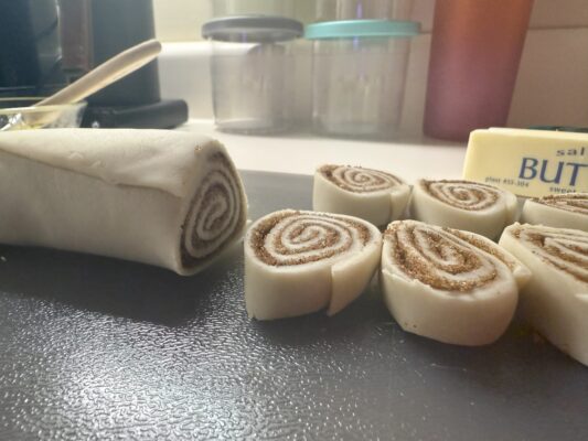 A roll of cinnamon and brown sugar between layers of dough sits next to sliced cinnamon rolls. In the background, there are Ninja Creami containers and a stick of butter.