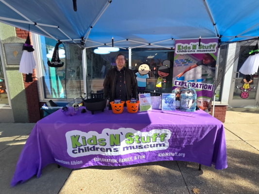 A man stands behind a table covered with a purple tablecloth. On the table are various toys and plastic Jack O’ Lantern bins. The front of the tablecloth reads “Kids ‘N’ Stuff children’s museum.”
