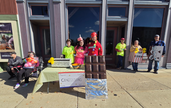 A wide shot shows many people standing or sitting outside a building with large glass windows, dressed as popular movie snacks with corresponding props surrounding their table.