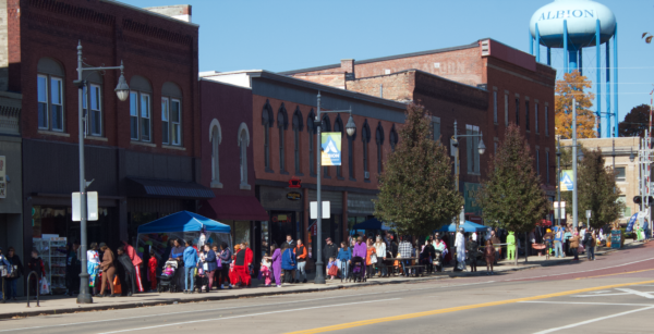 A line of families dressed up in colorful costumes stretches from the train tracks to the next intersection. Beside them are a row of buildings, with some tents set up on the sidewalk.