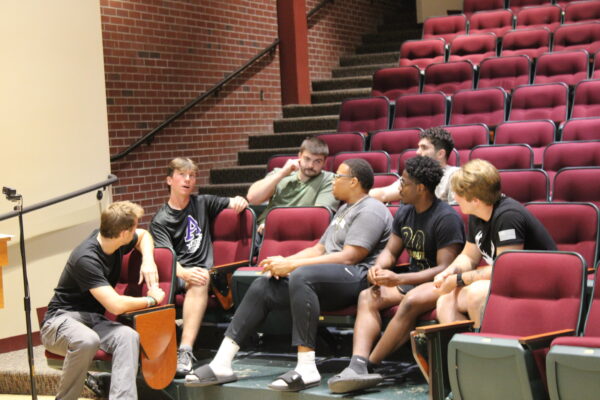 A group of young men sit in three rows of chairs in an auditorium talking to each other.