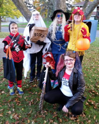 Five young pre-teens stand and crouch in front of a tree, smiling at the camera. A young boy is dressed as a ninja on the far left, a young girl as Harley Quinn in front, and two young people in the center as a ghost and a scarecrow, the person on the far right wears red and yellow with red mittens to look like crab claws and a crab hat, as well as a large blue cardboard shell. The photo is slightly blurry, and dead leaves are visible on the surrounding ground.