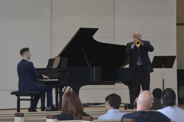An older man in a black suit and pale blue shirt on a stage plays the trumpet with the bell facing the camera. In the foreground are the heads and shoulders of several members of the audience. Behind him is a grand piano, where a man in a navy suit plays against a white background.