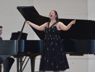 A young woman wearing a formal black dress with silver accents sings, head back and arms open. Behind her is a grand piano, where a man in a navy suit plays against a white background.