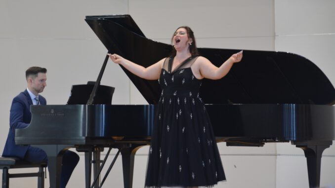 FEATURE A young woman wearing a formal black dress with silver accents sings, head back and arms open. Behind her is a grand piano, where a man in a navy suit plays against a white background.
