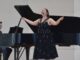 A young woman wearing a formal black dress with silver accents sings, head back and arms open. Behind her is a grand piano, where a man in a navy suit plays against a white background.