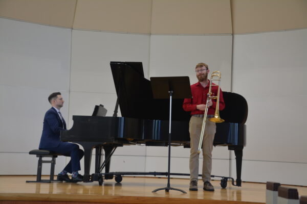 A young man in khaki pants and a red shirt stands on a stage holding a trombone and looking at a music stand in front of him. Behind him is a grand piano, where a man in a navy suit plays against a white background.
