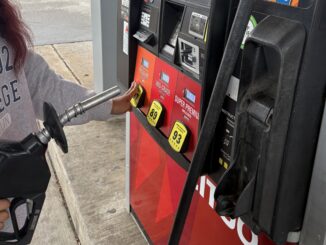 A gas station pump colored with red and orange decal stands at the center of the image, with different labels for ’regular,’ ‘mid-grade‘ and ‘super-premium’ gas. A metal pump is held up in frame, whilst a hand in a gray sweatshirt reaches out to select one of the fuel options.
