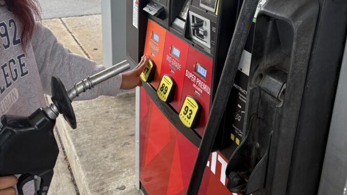 FEATURE A gas station pump colored with red and orange decal stands at the center of the image, with different labels for ’regular,’ ‘mid-grade‘ and ‘super-premium’ gas. A metal pump is held up in frame, whilst a hand in a gray sweatshirt reaches out to select one of the fuel options.