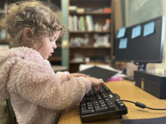 A young girl turned to the side types on a black computer keyboard. The girl is wearing a light pink fuzzy sweater and has her hair up in a bun. The background is a computer monitor with four blue sticky notes.