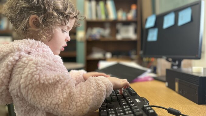 A young girl turned to the side types on a black computer keyboard. The girl is wearing a light pink fuzzy sweater and has her hair up in a bun. The background is a computer monitor with four blue sticky notes.