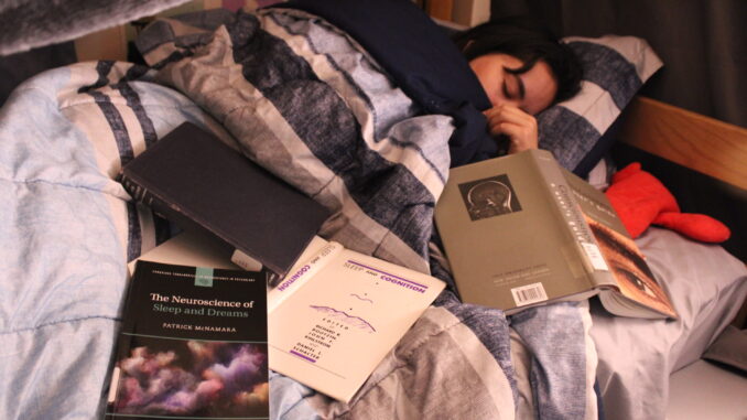 FEATURE A young woman with dark hair lays in a bed with light blue striped covers and a pillow under her head. Her eyes are closed, and she holds the covers up over her face. On the surrounding bed are four books, one in focus with the cover visible reading “The Neuroscience of Sleep and Dreams.”