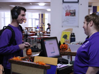 A young man wearing headphones, a purple crewneck and a black backpack smiles across a counter at an older woman wearing a purple collared shirt and a ribbon in her hair. On the counter between them are several Halloween decorations and a computer.