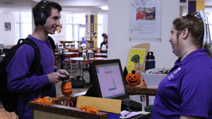 FEATURE A young man wearing headphones, a purple crewneck and a black backpack smiles across a counter at an older woman wearing a purple collared shirt and a ribbon in her hair. On the counter between them are several Halloween decorations and a computer.