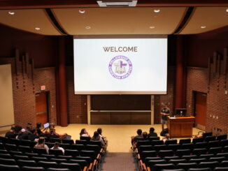 Young adults gather in the first rows of a lecture hall talking to one another. A screen attached to the ceiling has “welcome” written with a purple and gold version of the Fellowship of Christian Athletes logo.