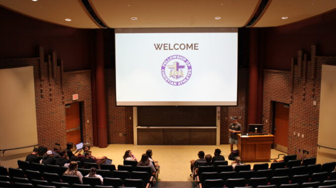 FEATURE Young adults gather in the first rows of a lecture hall talking to one another. A screen attached to the ceiling has “welcome” written with a purple and gold version of the Fellowship of Christian Athletes logo.
