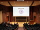 Young adults gather in the first rows of a lecture hall talking to one another. A screen attached to the ceiling has “welcome” written with a purple and gold version of the Fellowship of Christian Athletes logo.