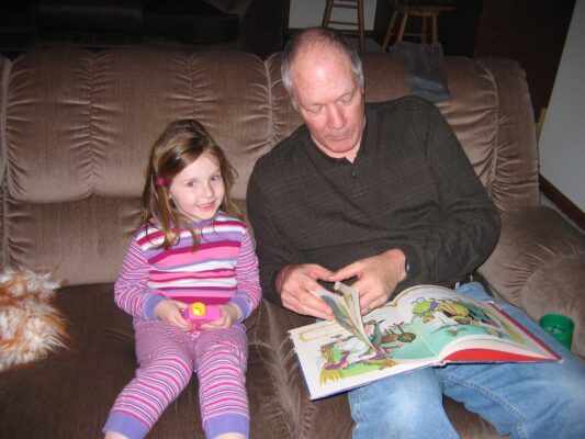 A young girl in striped pajamas sits on a brown couch and smiles as an old man reads her a picture book. 