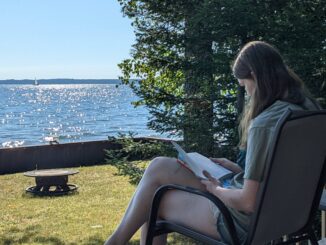 A young woman sits in a chair and reads a book. In front of the woman is a yard with a fire pit, a lake with the sun shining on it, pine trees and a blue sky.