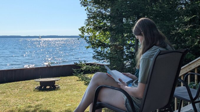 A young woman sits in a chair and reads a book. In front of the woman is a yard with a fire pit, a lake with the sun shining on it, pine trees and a blue sky.