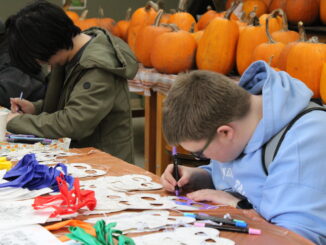 Two young individuals are crafting at a table covered with colorful masks and markers. In the background, lots of pumpkins create an autumnal setting.