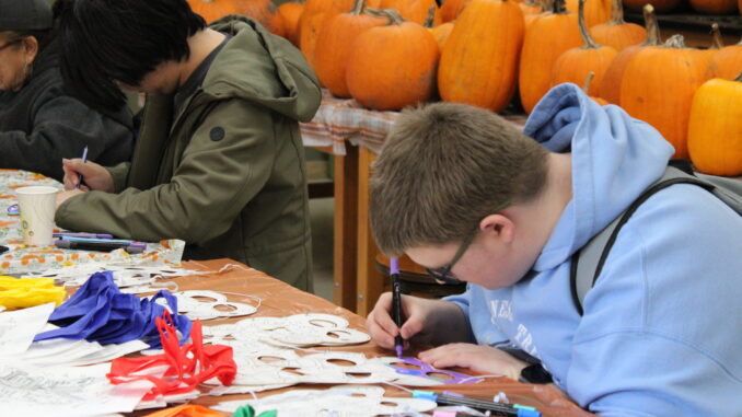 FEATURE Two young individuals are crafting at a table covered with colorful masks and markers. In the background, lots of pumpkins create an autumnal setting.