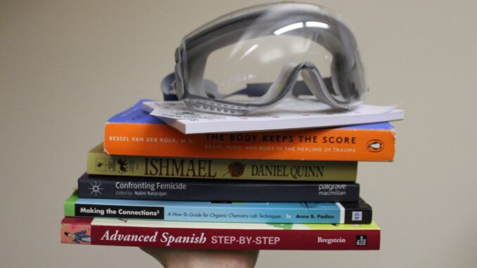 A series of books and objects are held up by one hand. From bottom to top are a Spanish textbook, chemistry lab guide, collection of essays on femicide, the book “Ishmael,” the book “The Body Keeps the Score,” a suicide prevention notepad and a pair of lab goggles.