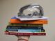 A series of books and objects are held up by one hand. From bottom to top are a Spanish textbook, chemistry lab guide, collection of essays on femicide, the book “Ishmael,” the book “The Body Keeps the Score,” a suicide prevention notepad and a pair of lab goggles.