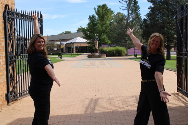 Two middle-aged women stand turned inward towards each other, their arms outstretched diagonally, smiling at the camera. They stand on a brick path with an open gate behind them, with a building, trees and green space visible in the background.