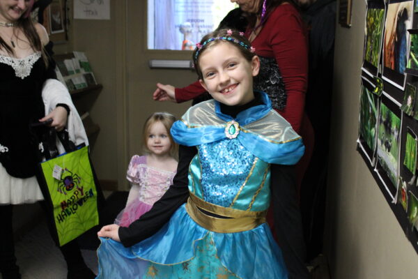 Two children in princess costumes stand in a hallway with adults in the background. In the foreground is a girl in blue smiling at the camera, with a younger child in pink behind her.