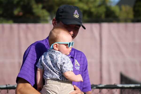 A man in a purple polo and black cap holds a baby wearing blue sunglasses and a floral shirt. They stand outdoors. 
