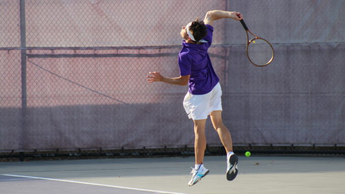 A tennis player in a purple shirt and white shorts leaps midair to serve. The scene is energetic, on a sunny day, with a chain-link fence backdrop.