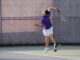 A tennis player in a purple shirt and white shorts leaps midair to serve. The scene is energetic, on a sunny day, with a chain-link fence backdrop.
