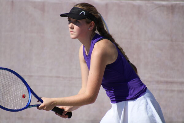 A focused female tennis player prepares to return a serve. She wears a purple tank top, white skirt, and black visor, holding a blue tennis racket. 