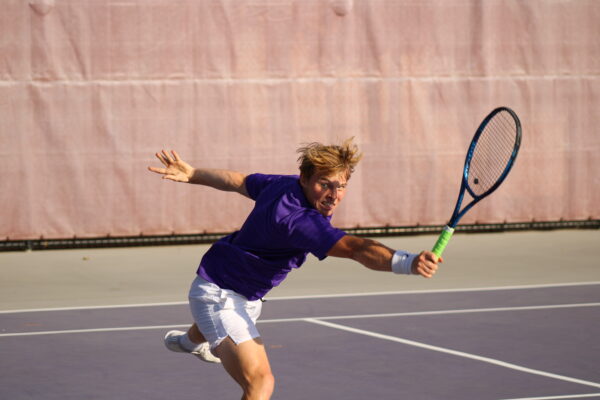 A tennis player in a purple shirt and white shorts lunges forward, swinging a racket with intensity. The background shows a tennis court and a pink canvas.
