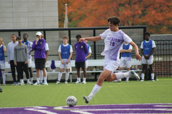 A soccer player in a white Albion uniform prepares to kick a ball on a field. Teammates in blue vests watch from the sidelines. Autumn trees in the background.