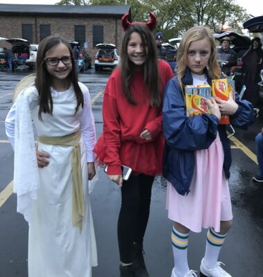 Three girls stand in a row in a parking lot. From left to right, they are dressed as an angel in a long white dress, a devil in red horns and a red tutu and a pink dress, blue jacket and tall socks holding boxes of Eggo waffles.