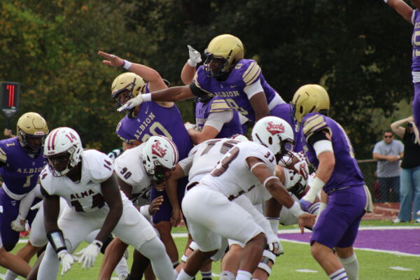 A football player in a purple Albion jersey leaps over teammates, attempting to score against opponents in white Alma jerseys on a grassy field, conveying determination.