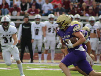 A football player in a purple Albion uniform runs with the ball, pursued by Alma opponents in white. The crowd watches, creating a tense, action-packed scene.