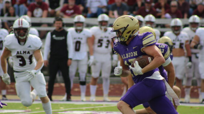FEATURE A football player in a purple Albion uniform runs with the ball, pursued by Alma opponents in white. The crowd watches, creating a tense, action-packed scene.