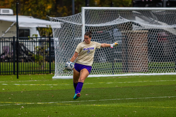 A soccer player in an Albion jersey kicks a ball on a green field, near a net. She displays focus and determination under a partly cloudy sky.